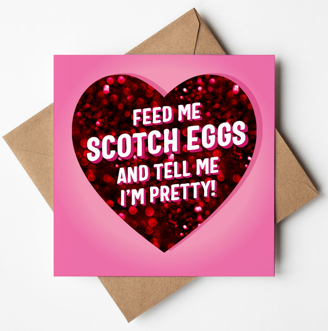 A funny card with a pink background features a red heart and the text "Feed me scotch eggs and tell me I'm pretty!" accompanied by a brown envelope.